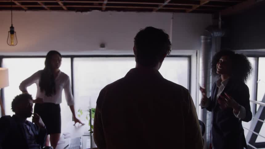 A group of young adult friends engage in a lively discussion indoors at midday gathered comfortably as sunlight filters through the windows highlighting their animated expressions and casual setting
