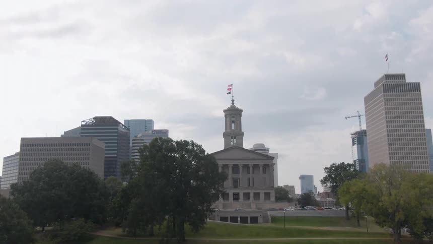 The video is an establishing shot of the tennessee state capitol building and the surrounding downtown nashville skyline on a cloudy day the state capitol stands prominently on a grassy hill framed by trees with modern highrise buildings in the
