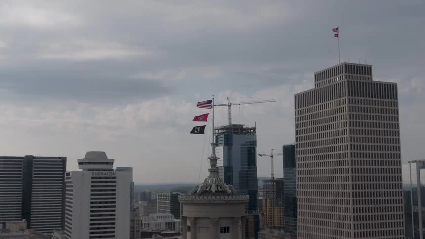 The video is an establishing shot of a city skyline against a cloudy sky focusing on two flags one american one black powmia flag waving on top of a tall building modern skyscrapers including one under construction with a crane visible sur