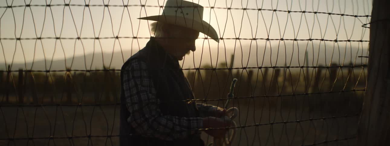 An old cowboy grabs a rope from a wire fence