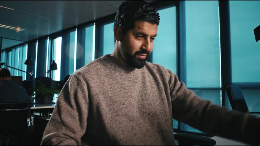 A man wearing a sweatshirt sits in an office at midday opening his laptop lid as he prepares to start work focused and ready to dive into the days tasks