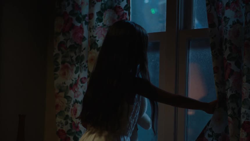 A girl pushes a curtain aside to look out a window at night