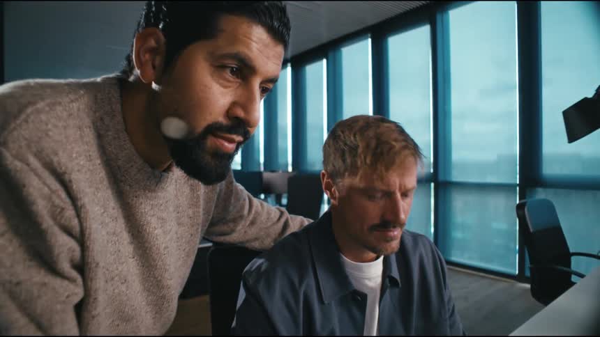 Two men work together on a computer in front of a screen in a corporate setting