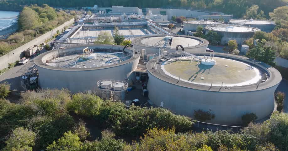 Reveal of circular tanks and other treatment facility buildings surrounded by trees and greenery