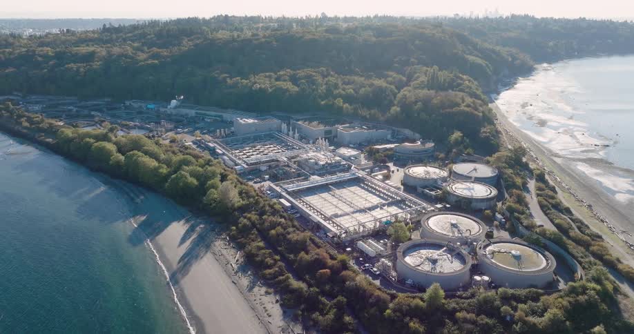 Sunlight reflects from a water treatment facility stretching between green forest and a tranquil bay with a city skyline in the distance