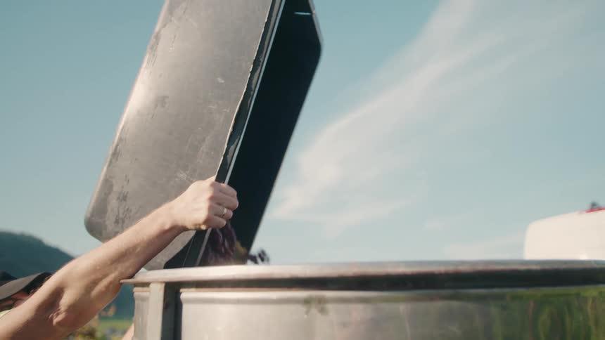 A man lifting a large black tray to dump grapes into a steaming silvercolored metal vat outdoors under a bright sky