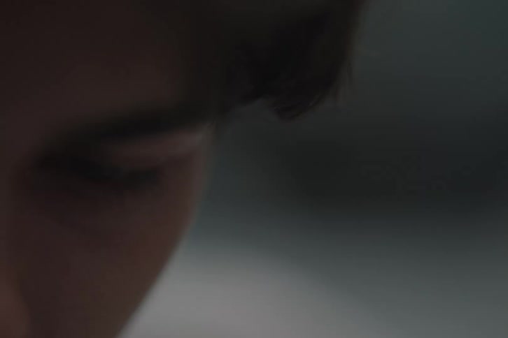 An extremely closeup shot focuses on the left eye and eyebrow area of a persons face with the eye blinking once and the gaze directed downward against a dark and completely outoffocus background