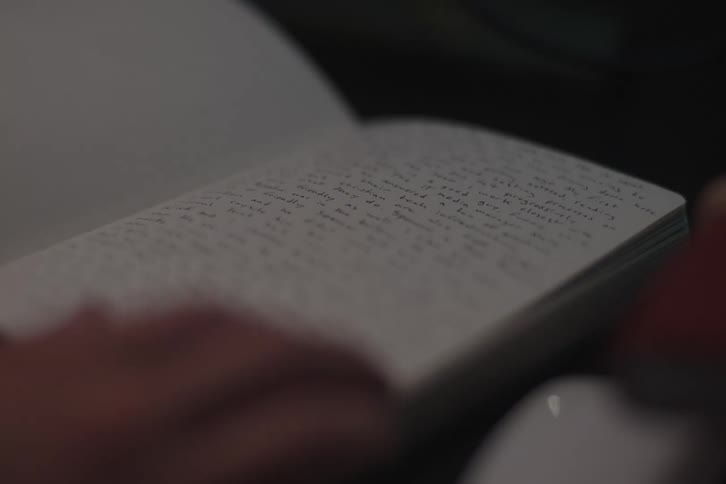 A close low angle shot of a persons hands turning pages in a darkened notebook filled with handwritten notes and text indicating private writing or study