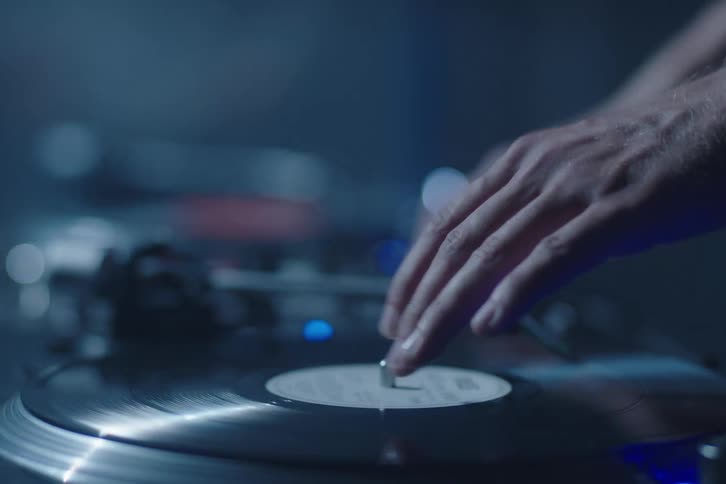 A close view shows the hands of a dj spinning and scratching a vinyl record on a turntable illuminated by blue and white party lights in a dark club environment