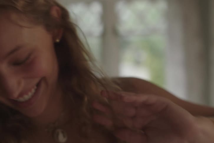 A blurry very close view of a smiling woman with long blonde hair wearing a white top laughs and looks down in a brightly backlit room