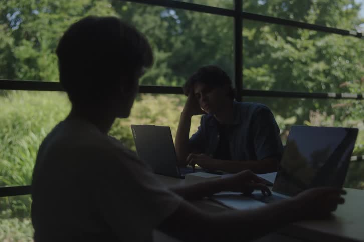 Two young men work on laptops near a bright window