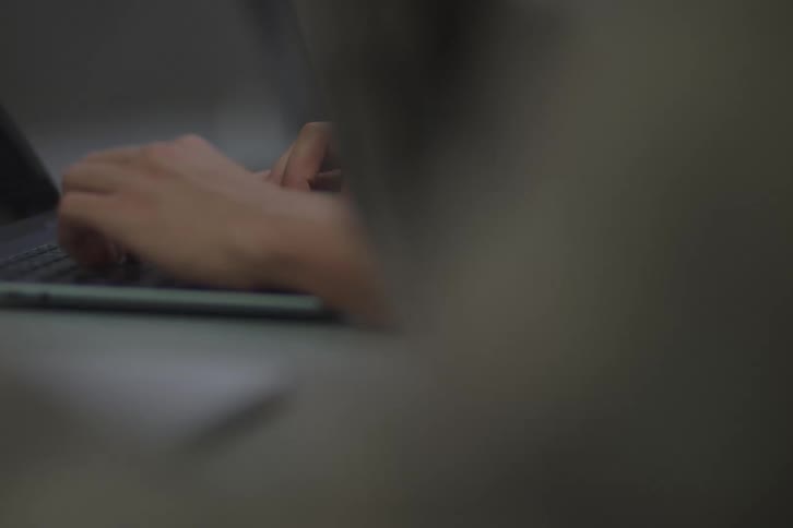 Male hands typing on a laptop computer with other people around also working on devices indoors during day time