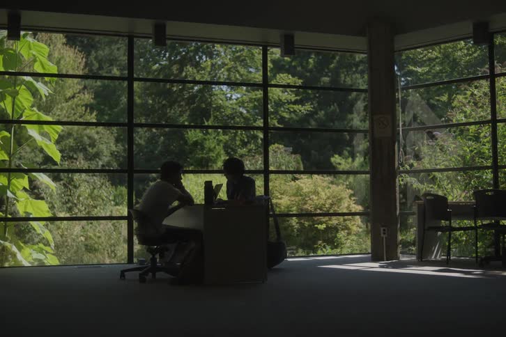 A low wide interior view shows a silhouette of a person walking in front of a brightly lit room where two people are sitting at a table with laptops all framed by large floor to ceiling windows overlooking a dense green forest
