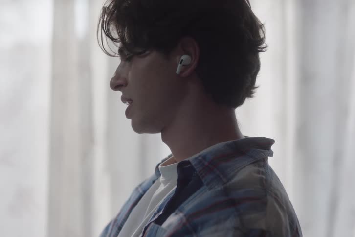 A young man with dark hair standing still wearing a wireless earbud in his visible ear with his shoulders and upper chest partially visible against curtains