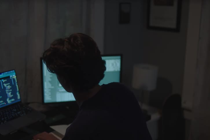 A teenage boy sitting in a dark room and looking intently at a large computer monitor displaying lines of code occasionally looking down at a smartphone held over an open notebook on a desk next to a glowing laptop screen