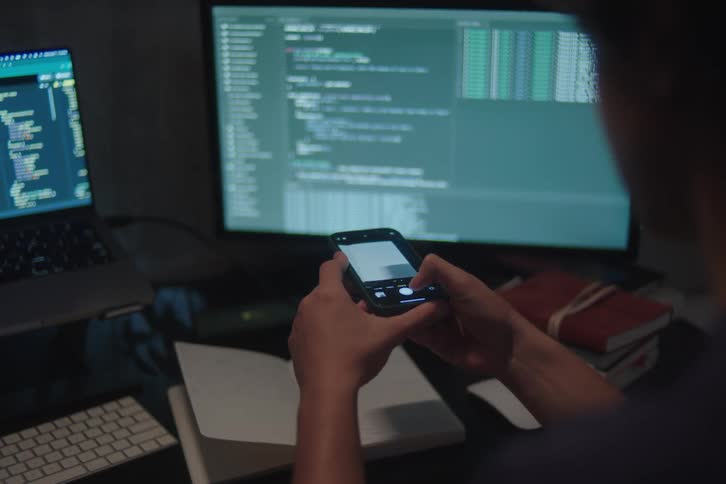 Person using a phone in front of monitors displaying programming code