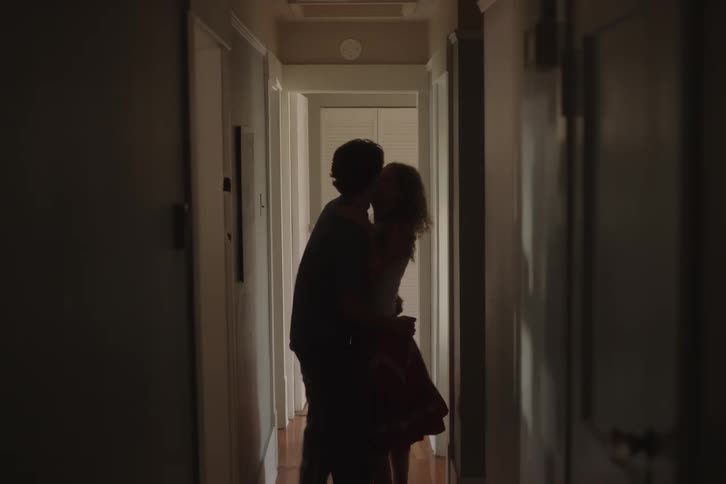 A couple dances and hugs in a dim hallway lit from a bright doorway with the woman in a red skirt and the man in dark overalls