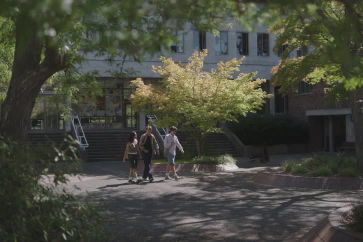 A long view through green trees shows three people in casual summer clothing walking toward the foreground on a paved outdoor path outside a large gray university building with steps and a bright yellow green tree in the center
