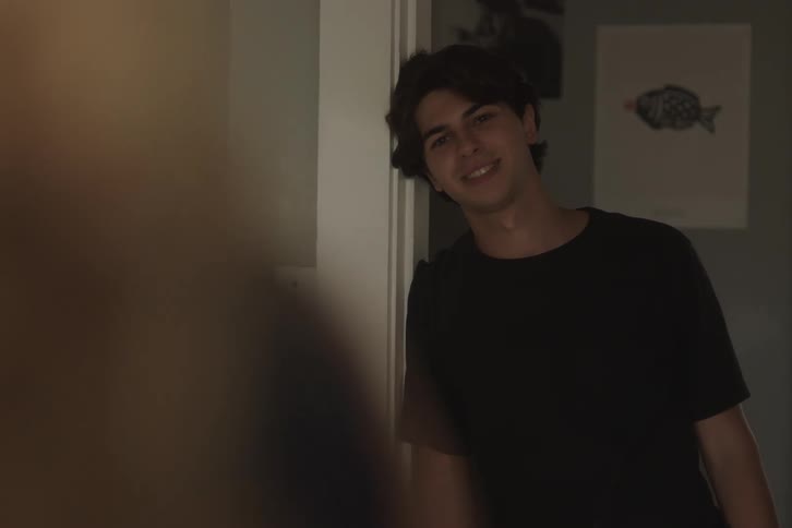 A young man with dark hair in a black shirt standing leaning against a doorframe and smiling at someone offcamera in a home setting