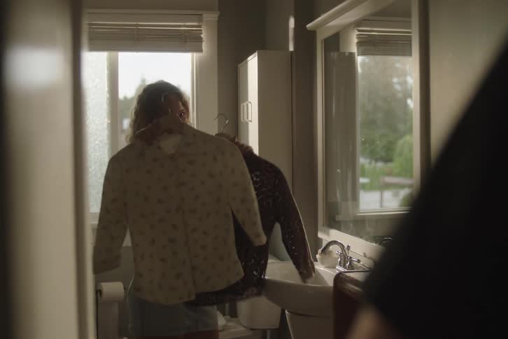 A young woman with curly hair standing next to a window in a bathroom briefly holding two articles of clothing while a mirror reflects the action and a sink and toilet are visible and theres a person in the foreground