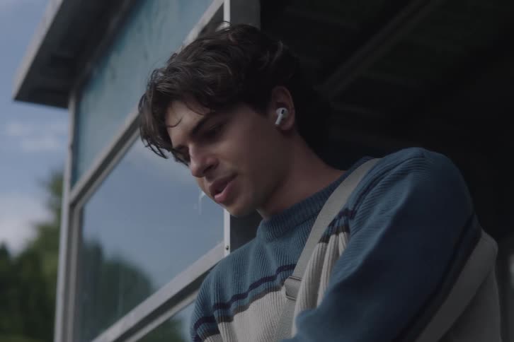 A young man wearing wireless earbuds leans against a windowed structure and looks out thoughtfully