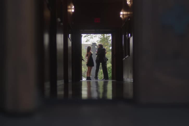A distant low angle view down a dark polished hallway shows a woman in a short dress and two men talking in the bright light of an open exterior doorway