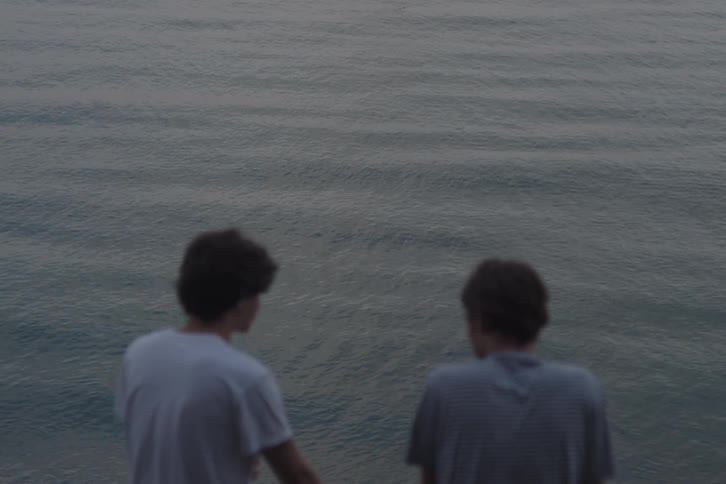 The backs and upper heads of two young mans standing and looking down at a large gray body of water with a consistent ripple pattern with the person on the left briefly turning their head toward the person on the right