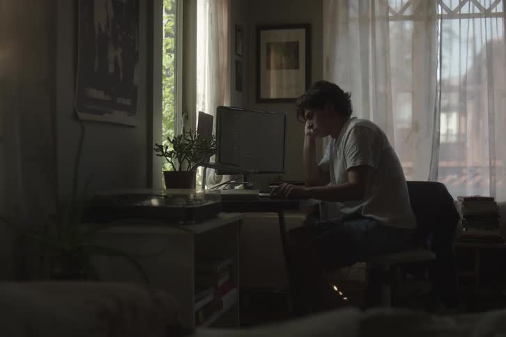 A young man wearing a lightcolored tshirt and dark pants sitting at a desk and looking at a computer monitor in a dimly lit room with bright natural light strongly backlighting the scene through a curtaincovered window
