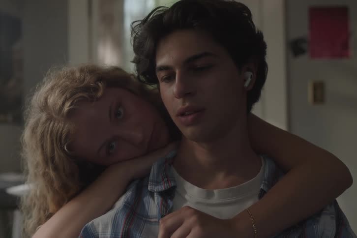 A young woman with long blonde hair and a white tank top embraces a young man with dark curly hair and white wireless earbuds from behind resting her head on his shoulder in a soft intimate moment