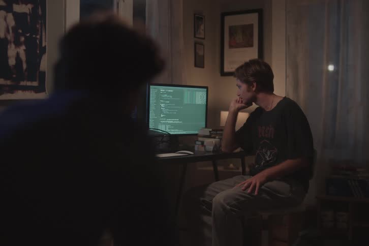 A young man sits in front of a computer monitor engaged in a focused discussion with another man beside him