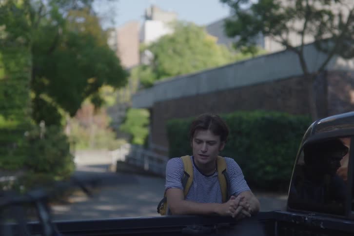 A low angle shot from the back of a truck shows two young men walking toward the camera on a paved path outside a university building with one pushing a bicycle and the other stopping to lean on the truck bed
