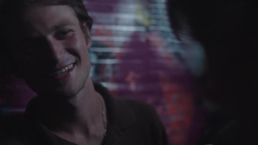 A dark close view of a smiling man with light brown hair and a dark polo shirt looking happily at someone off camera with colorful graffiti blurred in the background