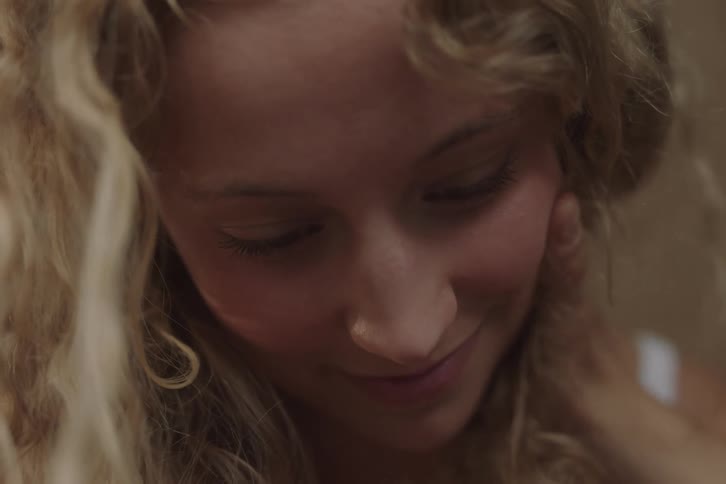 An extreme closeup shot focuses on the face of a woman with long curly hair who looks downward and shows a subtle smile while a second person out of focus touches her face