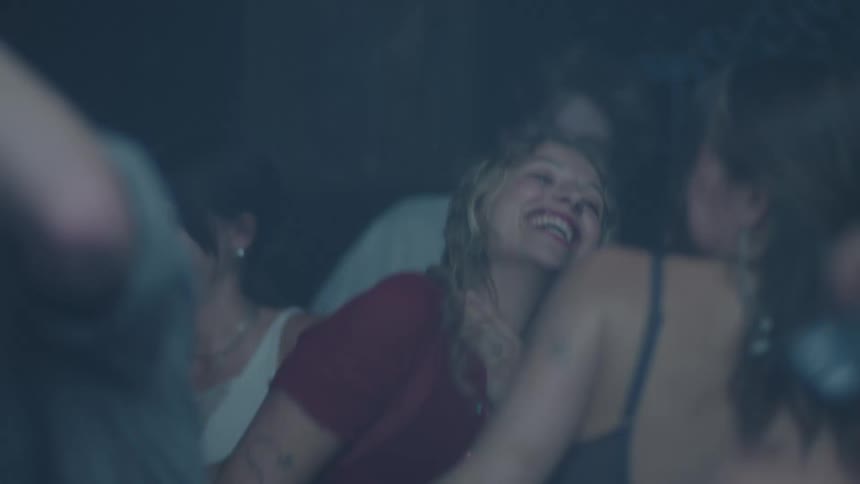 A medium close shaky view captures a group of smiling young people in dark clothing dancing wildly and laughing together under flashing blue lights in a dark club or party atmosphere