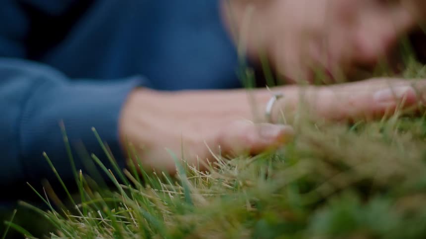 A woman lies quietly in the grass with a relaxed hand grazing the blades