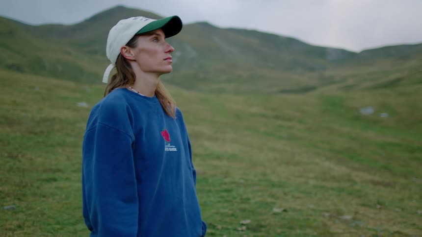 A woman in a sweatshirt stands on a grassy slope overlooking mountains