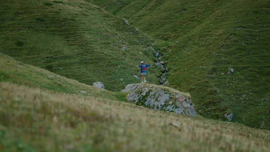 A person walks on a mossy rock with arms outstretched in a grassy mountain valley