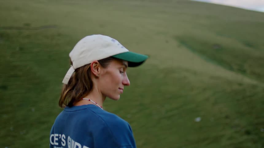 A young blonde woman wearing a baseball hat stands on a sunlit grassland at midday glancing around