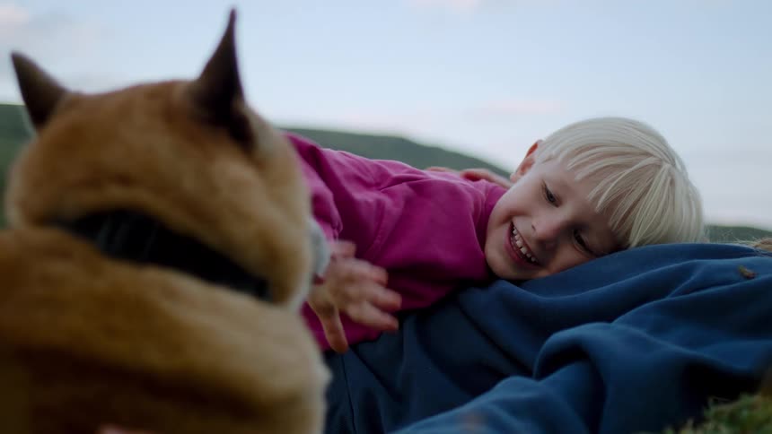 A smiling blond child in pink rests on a blue sleeved adult while a tan dog nuzzles close