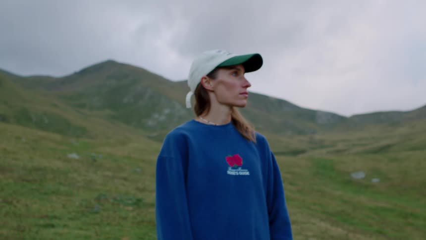 A woman in a blue sweatshirt and cap stands in a green mountain meadow looking upward with a calm thoughtful expression