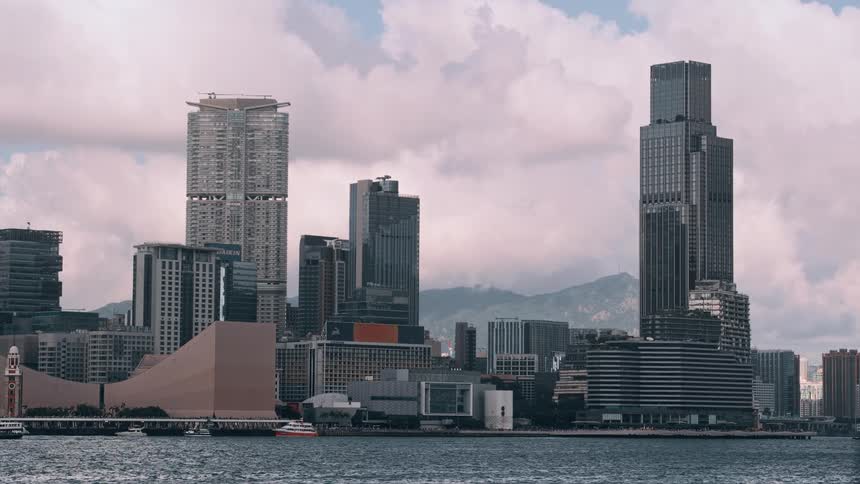 A wide view across calm water shows the dense skyline of a modern city composed of multiple tall skyscrapers and architectural buildings under a partly cloudy sky with mountains in the background