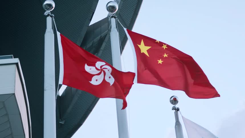 The flag of hong kong and china sidebyside both waving on tall white flagpoles against a bright sky and in front of an indistinct modern building structure