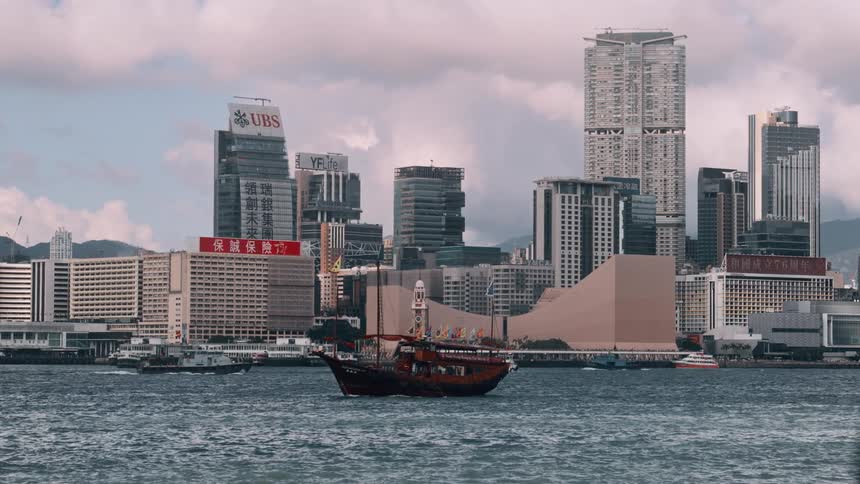 A traditional junk boat sailing past modern high rises under a cloudy sky