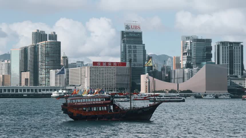 A tourist junk boat with colorful flags sails across the water against the backdrop of the modern hong kong skyline under a cloudy sky