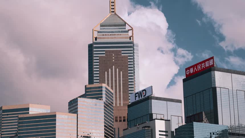 A city skyline shows modern glass skyscrapers with a tall tower capped by a gold triangular peak and a building displaying red chinese signage