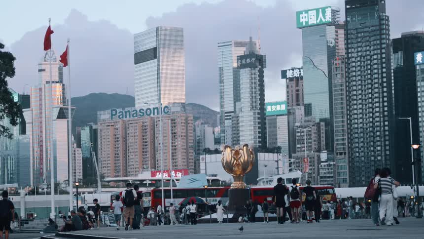 The video is a wide shot of a crowded modern city center featuring towering glass skyscrapers in the background in the foreground there are many people walking and sitting near a large golden cupshaped sculpture and a red doubledecker bus