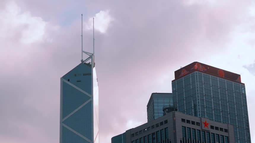 A low angle view of a modern city skyline shows the angular bank of china tower and another skyscraper with red chinese characters on top set against a pink and gray cloudy sky