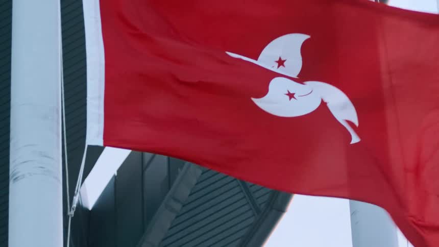 The hong kong flag flies beside a highrise building on a white pole blowing in the wind at midday