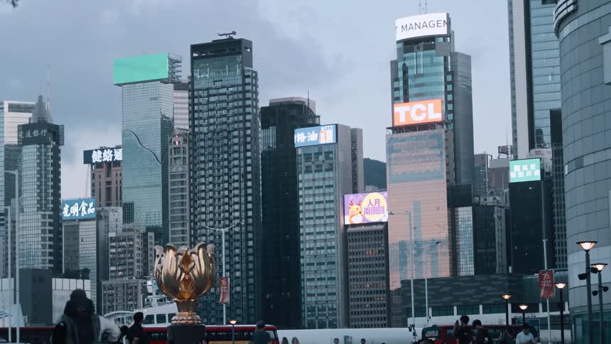 The video displays a dense array of skyscrapers in a city likely hong kong with various illuminated signs on their facades in the foreground there is a large golden cupshaped sculpture and the silhouettes of people and red doubledecker bus