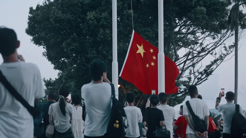 The video shows a crowd of people standing beneath two flagpoles at dusk watching the flags the central flag is the red chinese flag with five yellow stars and most of the people in the foreground are taking pictures with their phones